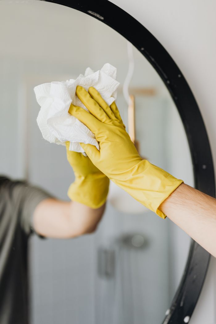 About Crop male in yellow latex glove wiping mirror with paper napkin and detergent in bathroom while standing and working at home